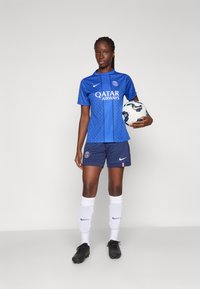 Blue soccer jersey with geometric pattern, white logo text, navy shorts, black cleats, and white socks. Player holds a soccer ball.