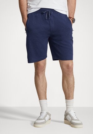 Mann trägt marineblaue Shorts mit Kordelzug, weiße Sportsocken, grau-weiße Turnschuhe und ein weißes T-Shirt, die Hände in den Taschen.