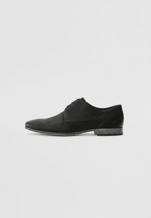 Zapato negro de vestir con puntera afilada, que presenta una textura estriada, acentos lisos y un tacón bajo. El diseño es elegante y moderno.