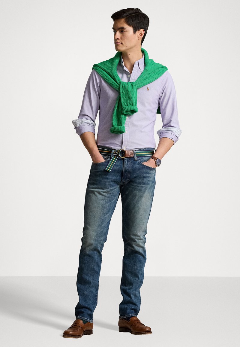 Lichtpaarse overhemd met knopen, groene gebreide sweater over de schouders, blauwe spijkerbroek, bruine leren schoenen en een multicolor gestreepte riem.