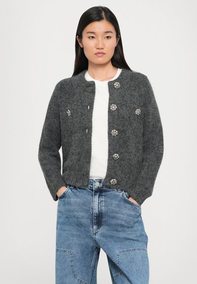 Harmaa villa cardigan, jossa on pyöreä kaula-aukko ja kuusi kukkakuvioista nappia. Yhdistetty valkoisen paidan kanssa ja vaaleansinisten farkkujen kanssa.