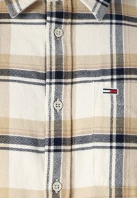 Camisa de franela a cuadros en tonos beige y crema con rayas azules y negras, que presenta un bolsillo en el pecho y cierre de botones. Textura suave, diseño clásico.