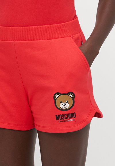Pantaloni corti rossi in cotone con grafica di un orso e testo "MOSCHINO UNDER BEAR". Vita elasticizzata e tasche laterali visibili.