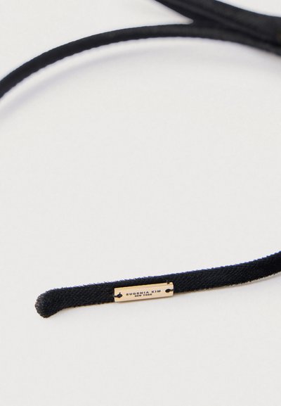 Elastic pentru păr din material negru, prevăzut cu o etichetă metalică aurie gravată cu „EUGENIA KIM NEW YORK.” Textură netedă, cu capete rotunjite.