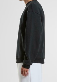 Personne portant un sweat-shirt noir ample à manches longues avec des poignets côtelés et un pantalon blanc, vue de côté sur un fond uni.