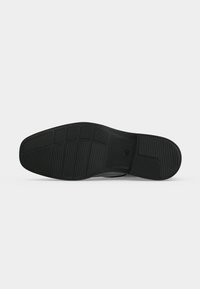 Semelle de chaussure noire avec des motifs de grip texturés incluant des sections rectangulaires et circulaires sur un fond gris uni.