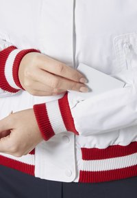 Bílá bomber bunda s vroubkovanými červenými a bílými manžetami, která má na rukávu zipovou kapsu na malé předměty.