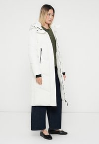 Kvinde iført lang hvid pufferjakke med hætte, grøn sweater, brede mørke jeans og sorte balletflade sko, stående mod en ensfarvet hvid baggrund.