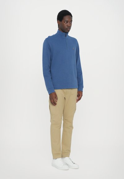 Blauwe zip-neck trui met lange mouwen, gecombineerd met beige cargobroeken en witte sneakers. De trui heeft een klein logo op de borst.