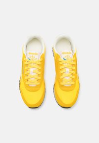 Scarpe da ginnastica gialle con tomaia in tessuto morbido, punta rotonda e design con lacci. Presentano un interno bianco e una suola in gomma nera con accenti.