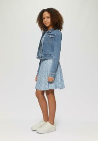 Denimjacke mit vorderen Knöpfen, kurzen Ärmeln, kombiniert mit einem hellblauen Blumenkleid. Weiße Sneakers runden das Outfit ab.
