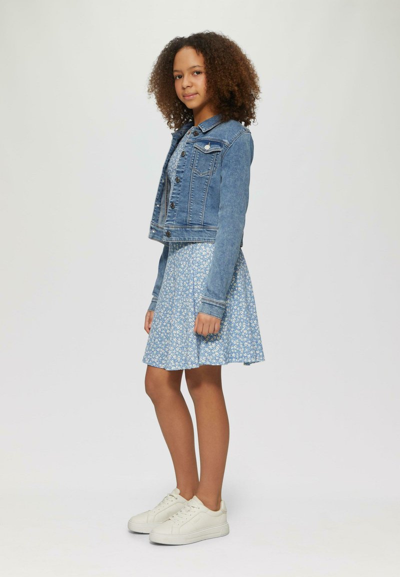 Denimjacke mit vorderen Knöpfen, kurzen Ärmeln, kombiniert mit einem hellblauen Blumenkleid. Weiße Sneakers runden das Outfit ab.