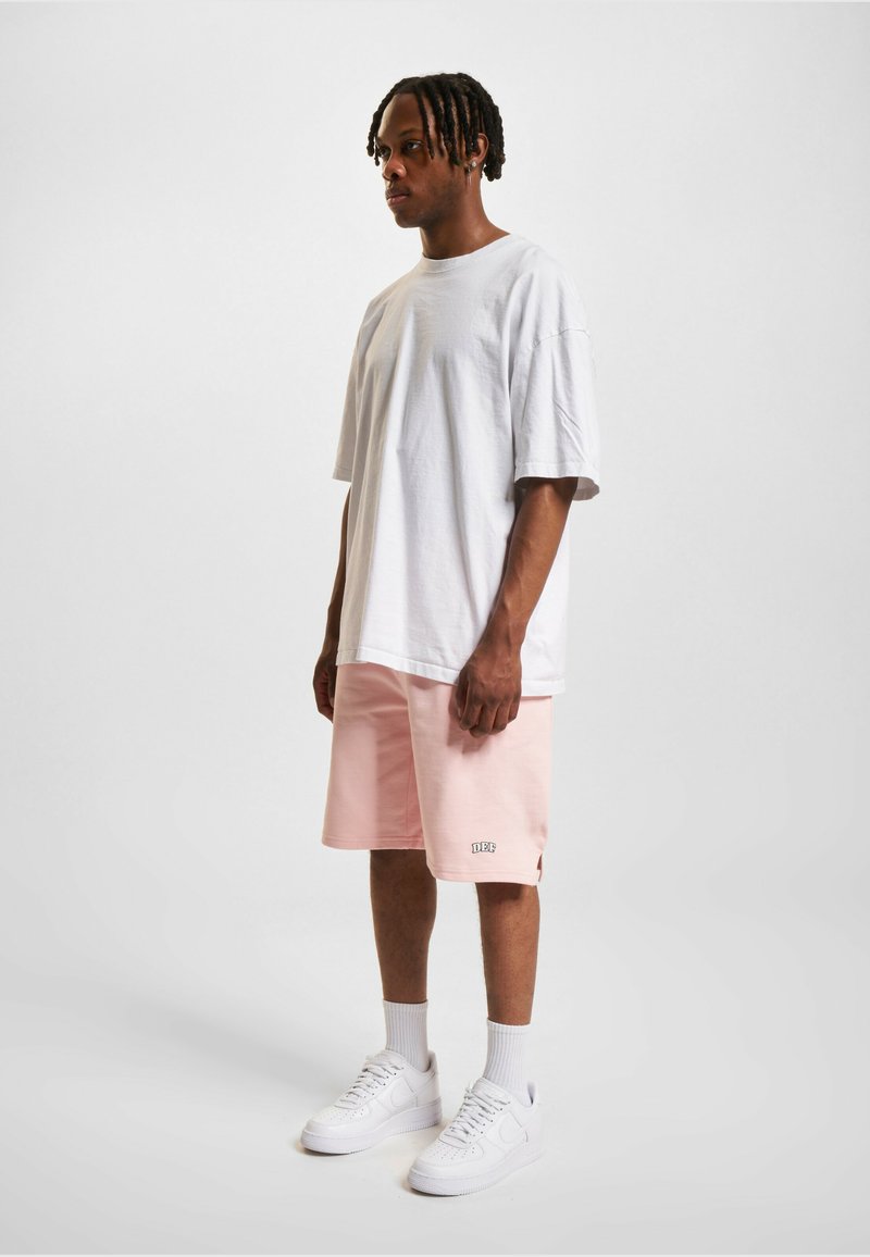 T-shirt oversize blanche, shorts roses avec logo, et baskets blanches. Texture lisse, coupe décontractée, shorts longueur genoux.