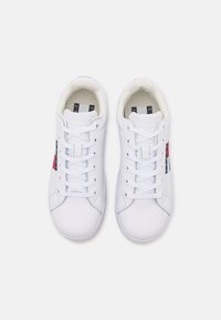 Baskets synthétiques blanches avec une texture lisse, des lacets plats et un patch logo avec des accents rouges et bleus sur le côté extérieur.