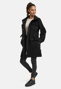 Cappotto nero con zip frontale, quattro tasche, vita regolabile e cappuccio. Realizzato in tessuto morbido e testurizzato. Abbinato a jeans neri e sneaker.