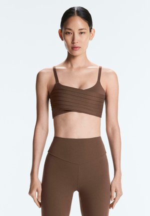 Kobieta stojąca, ubrana w brązowy, prążkowany krótki top na cienkich ramiączkach i pasujące legginsy z wysokim stanem na jednolitym jasnym tle.