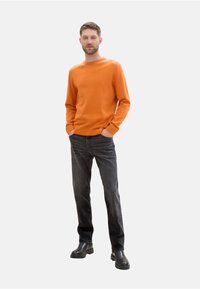 Jersey de punto naranja con mangas largas, cuello redondo, combinado con jeans oscuros y botas negras hasta el tobillo, de pie sobre un fondo blanco.
