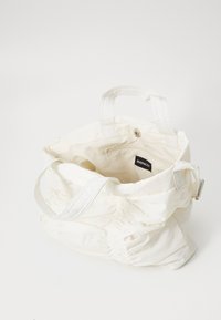 Witte canvas draagtas met geplooide textuur, twee stevige handvatten en een knoopsluiting. Binnenzak heeft een merketiket.