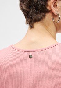 Ροζ ριμπάτ τοπ με στρογγυλή λαιμόκοψη και μια μικρή διακοσμητική πινέζα στην πλάτη. Το ύφασμα φαίνεται μαλακό και υφασμένο.