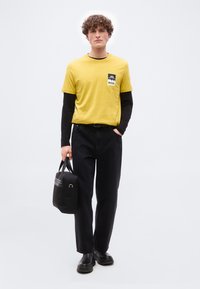 Jeune homme aux cheveux bouclés portant une chemise jaune à manches courtes par-dessus un sous-pull noir à manches longues, un pantalon noir, des chaussures noires, tenant un sac noir.