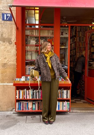 Vrouw in een luipaardjas en gele sjaal staat buiten bij een rood boekenwinkel met boekenkasten, houdt een groene handtas vast en kijkt naar de zijkant.