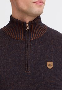 Granatowy sweter z dzianiny z wysokim prążkowanym kołnierzem, brązowym suwakiem oraz beżową naszywką z logo misia. Miękka struktura z subtelnymi plamkami.