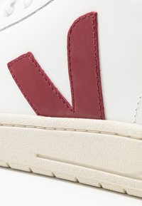 Zapatilla blanca con un gran logotipo rojo, suela texturizada de color blanco roto y parte superior de cuero suave. Presenta costuras en contraste y un diseño minimalista.