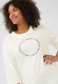 Camiseta de manga larga color crema con un diseño gráfico de texto redondo en negro, con escote redondeado y corte relajado.