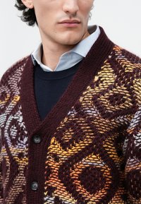 Πλεκτό cardigan σε σκούρο μπορντό με περίπλοκα κίτρινα και γκρι σχέδια, διαθέτοντας V λαιμόκοψη, κουμπιά και φορεμένο πάνω από ένα ναυτικό πουλόβερ.