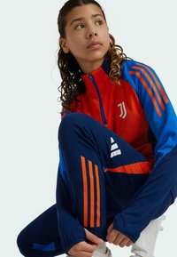 Juventus Trainingsanzug in Rot, Blau und Marineblau; mit orangefarbenen Streifen, Reißverschlussjacke und passenden Hosen mit gestreiftem Design und Logo-Akzenten.
