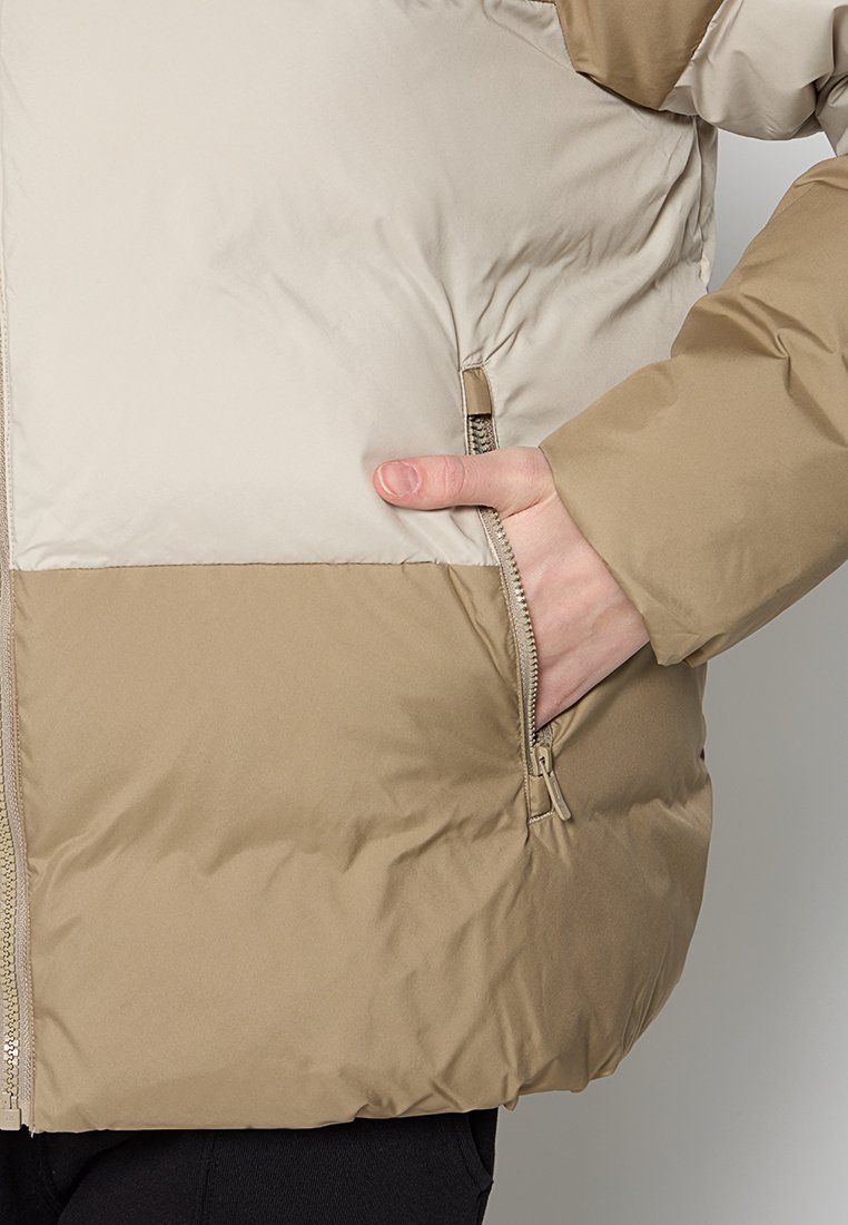 Ręka wsunięta do zamykanej kieszeni beżowej i jasnobeżowej kurtki puchowej z widocznym przeszyciem i teksturą materiału.