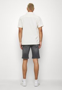 Camicia a maniche corte beige chiaro con colletto, abbinata a pantaloni corti in denim scuri. Il modello indossa scarpe da ginnastica bianche e si trova davanti a uno sfondo neutro.