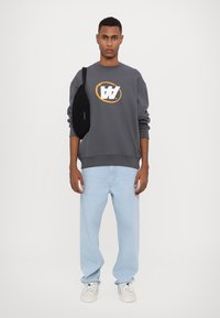 Grijze sweater met een geel en wit logo, gecombineerd met lichtblauwe jeans en witte sneakers. Het model houdt een zwarte schoudertas vast.