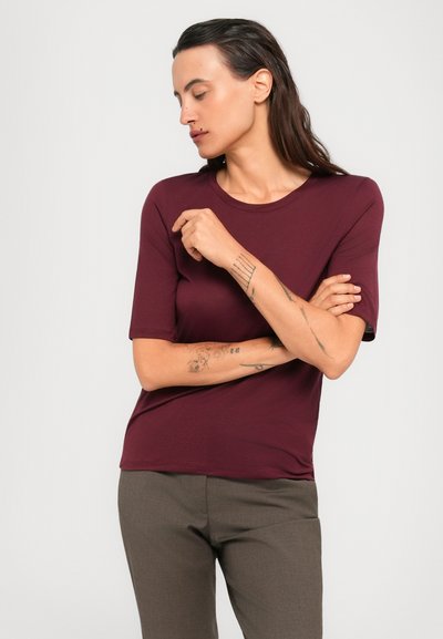 Woman with long dark hair, wearing a burgundy short-sleeve top and gray pants, crosses arms and looks downward against a plain background.