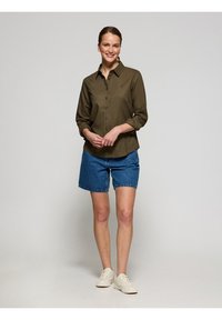 Olivgrön skjorta med knappar och trekvartslång ärm, ihopparad med denimshorts. Modellen bär vita sneakers; neutral bakgrund.