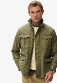 Mand iført en grøn knapjakke over en ternet skjorte og en beige sweater, stående med den ene hånd i lommen, hvid baggrund.