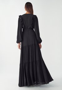 Black maxi dress with ruffled sleeves, a fitted bodice, and tiered skirt. Made of smooth fabric, featuring a back zipper and a waist tie.