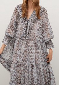 Nainen, joka on pukeutunut pitkähihhaiseen, laskostettuun, kerrokselliseen mekkoon, jossa on violetteja, ruskeita ja harmaita abstrakteja kuvioita sekä röyhelökaulusyksityiskohtia.