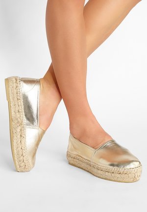 Piernas cruzadas con zapatos tipo espadrilles metálicos dorados sin cordones con suelas plataformas gruesas de yute trenzado sobre fondo blanco.