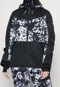 Kvinna iklädd en svart- och vitblommig mönstrad vinterjacka med matchande byxor, dragkedja framtill och tumhål i ärmarna.