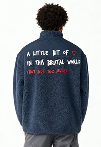 Veste polaire bleu marine avec un col haut, une texture douce, et un texte blanc et rouge au dos : « Un peu d'amour dans ce monde brutal. »