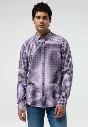 Mężczyzna w czerwono-biało-niebieską kraciastą koszulę na guziki i niebieskie dżinsy, stojący na tle jednolitego jasnego tła.