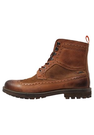 Stivaletto alla caviglia in pelle marrone con design wingtip brogue, occhielli metallici, chiusura frontale con lacci, suola in gomma e linguetta posteriore per facilitare la calzata.