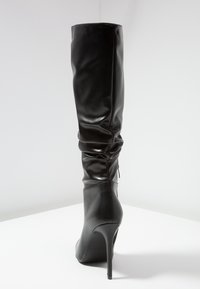Bota negra por encima de la rodilla hecha de material sintético suave, con punta afilada, tacón alto de agujas y detalle fruncido en la parte superior.