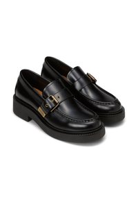 Mocassins en cuir noir avec un design piqué, présentant une boucle décorative et une semelle noire épaisse et texturée.