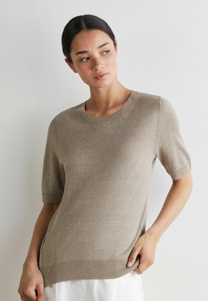 Frau mit dunklen Haaren, die einen beigen kurzärmligen Strickpullover und einen weißen Rock trägt, schaut leicht seitlich vor einer schlichten weißen Wand.