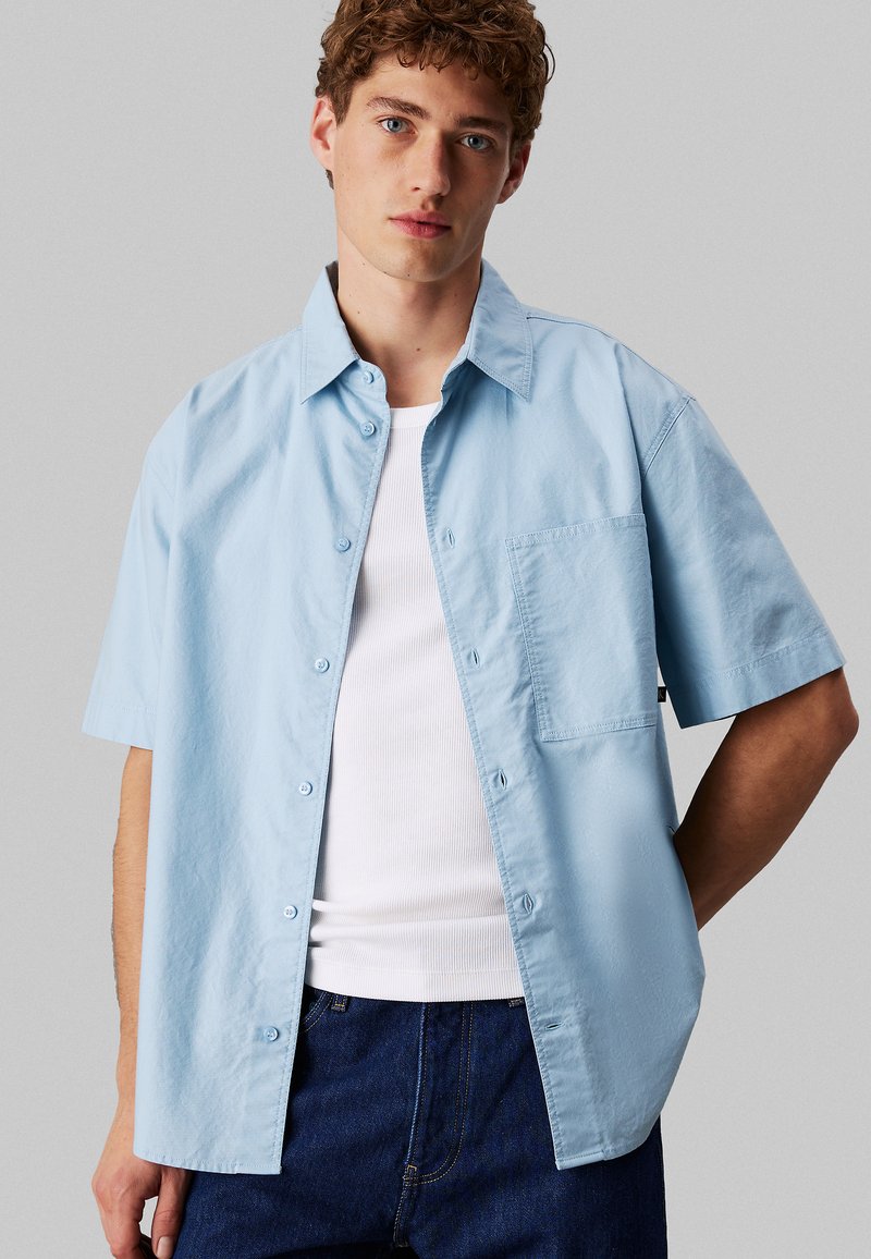 Camicia azzurro chiaro a maniche corte con design con bottoni, tasca singola sul petto, realizzata in tessuto morbido, abbinata a una canottiera bianca e jeans scuri.