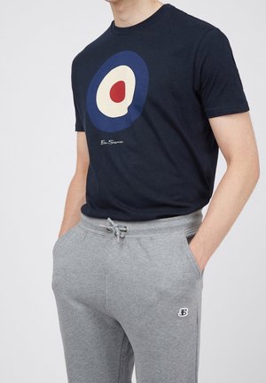 Mann trägt ein dunkelblaues T-Shirt mit einem kreisförmigen roten, weißen und blauen Design und graue Jogginghose mit Kordelzug und Logo auf der Tasche. Hände in den Taschen.