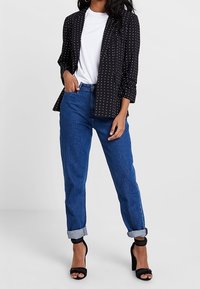 Blazer noir à pois blancs, t-shirt blanc, jean bleu retroussé, et sandales noires à talons hauts. Tenue décontractée avec une coupe contemporaine.