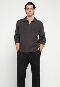 Barna kötött pulóver galléros nyakkivágással és három gombbal, bordázott mandzsettákkal és szegéllyel, fekete nadrággal párosítva.